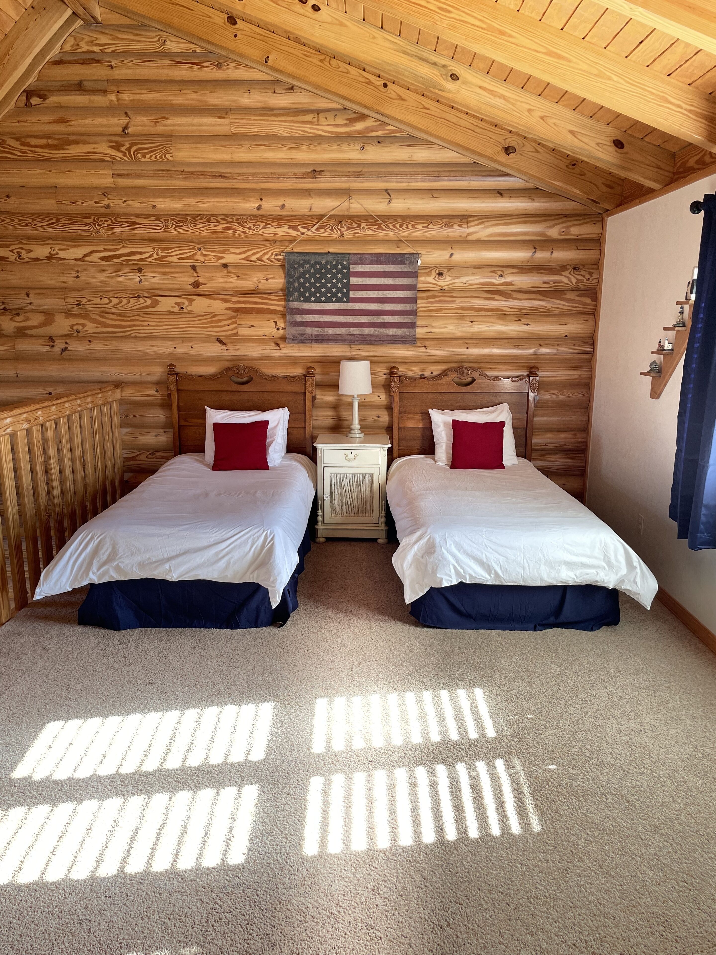 A room with two beds, wooden walls, a ceiling with wooden beams, a small nightstand with a lamp, and an American flag on the wall.