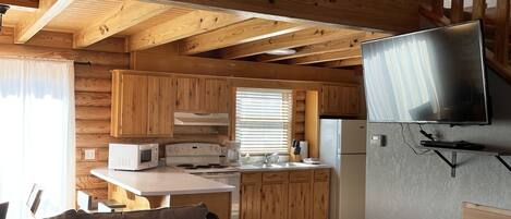A cozy cabin interior with a wooden staircase, a kitchen area with a microwave and stove, and a sofa with patterned cushions.