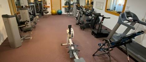 Un gimnasio con diversos equipos de ejercicio, incluyendo cintas de correr y máquinas de pesas, dispuesto en una sala amplia con alfombra roja.