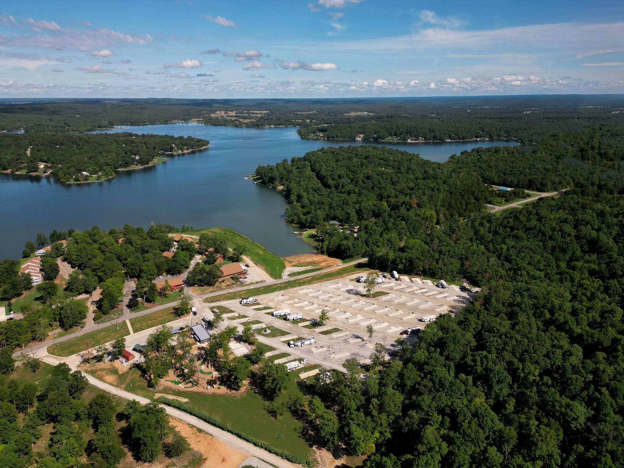 Vue aérienne d’un terrain de camping comprenant plusieurs aires de stationnement pour véhicules récréatifs, un lac et des zones boisées environnantes.