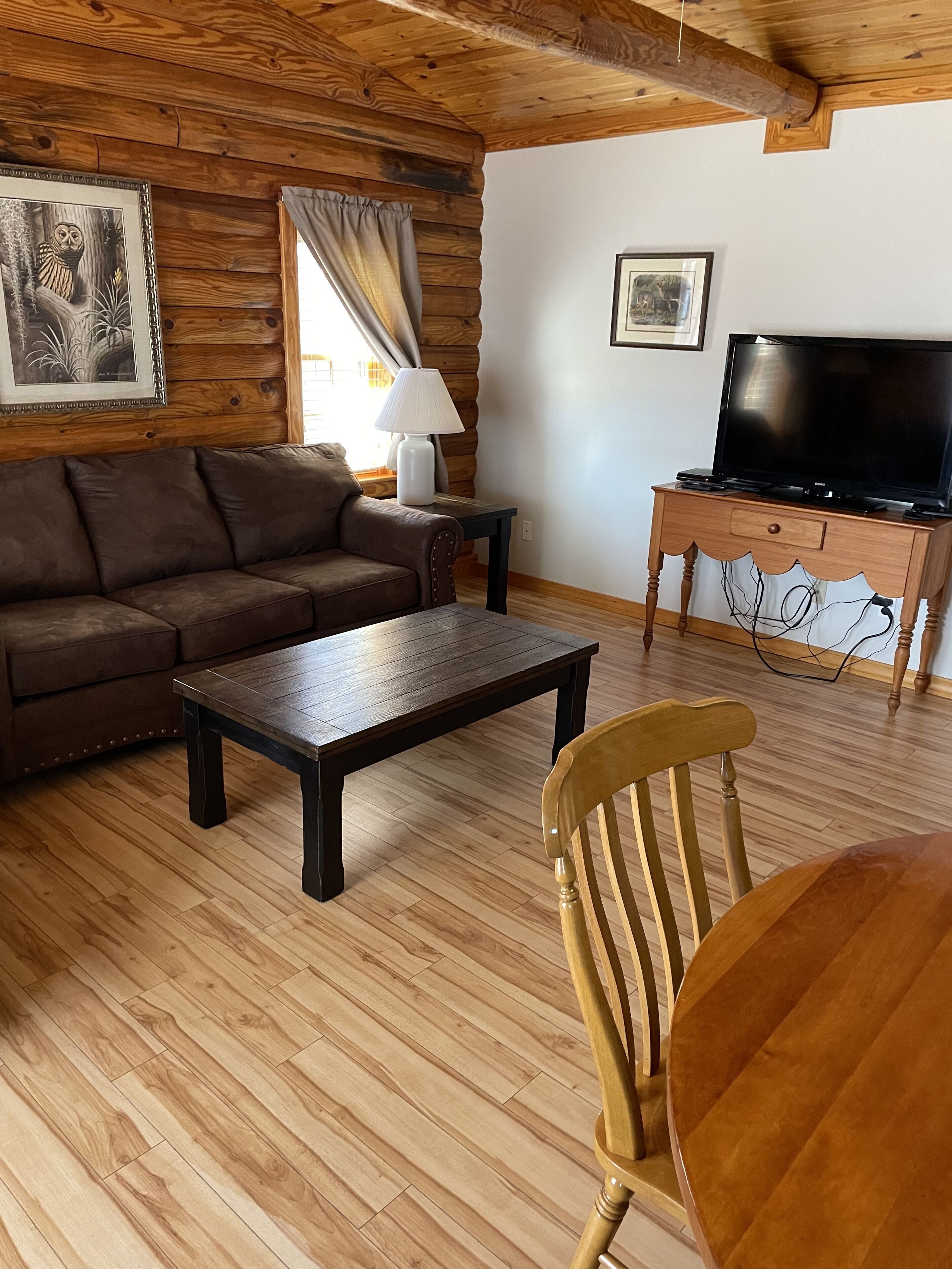 A cozy living room with a wooden ceiling, a brown sofa, a coffee table, a wooden chair, a TV on a wooden stand, and a lamp on a small table.