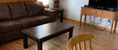 A cozy living room with a wooden ceiling, a brown sofa, a coffee table, a wooden chair, a TV on a wooden stand, and a lamp on a small table.
