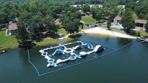 An aerial view of a lakeside park with various water play structures, a sandy beach, and surrounding residential houses.