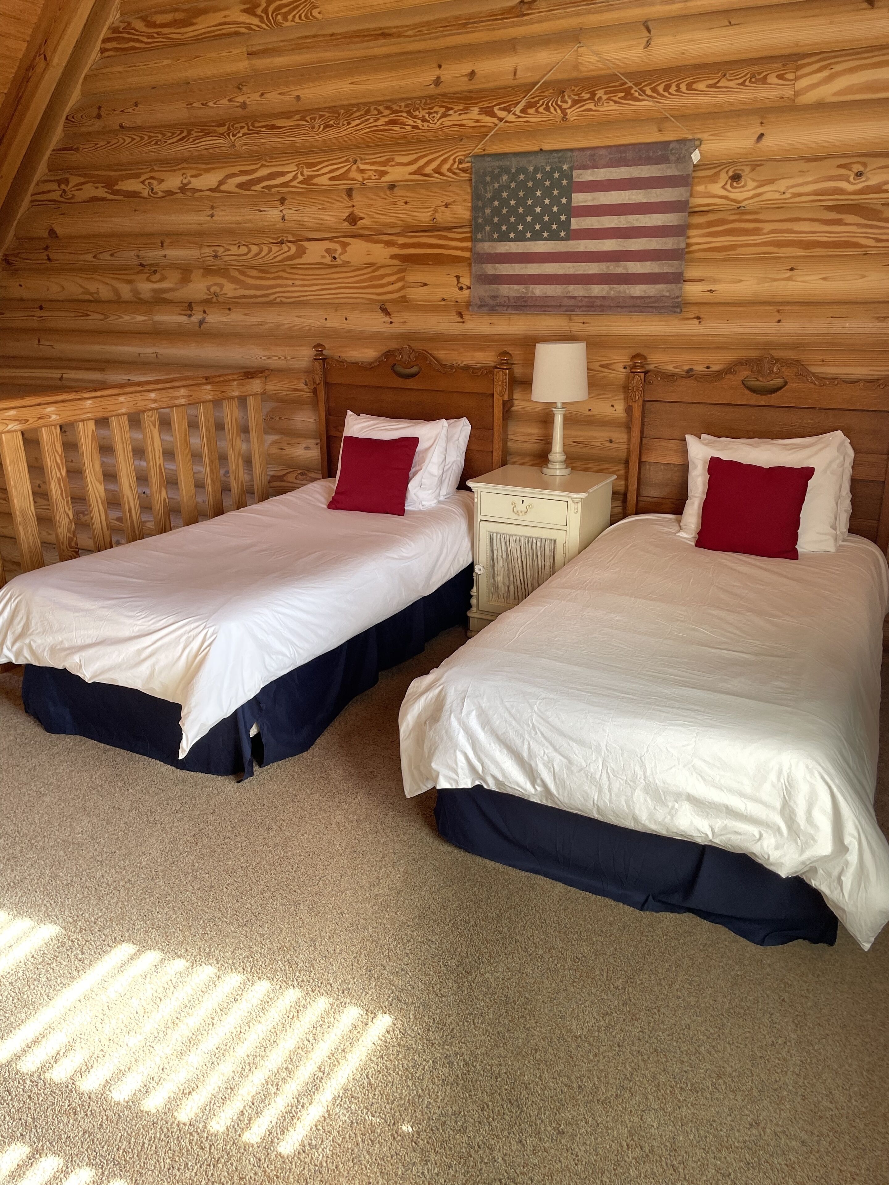A cabin with two beds, each with white bedding and red pillows, wooden walls, and a small nightstand with a lamp.