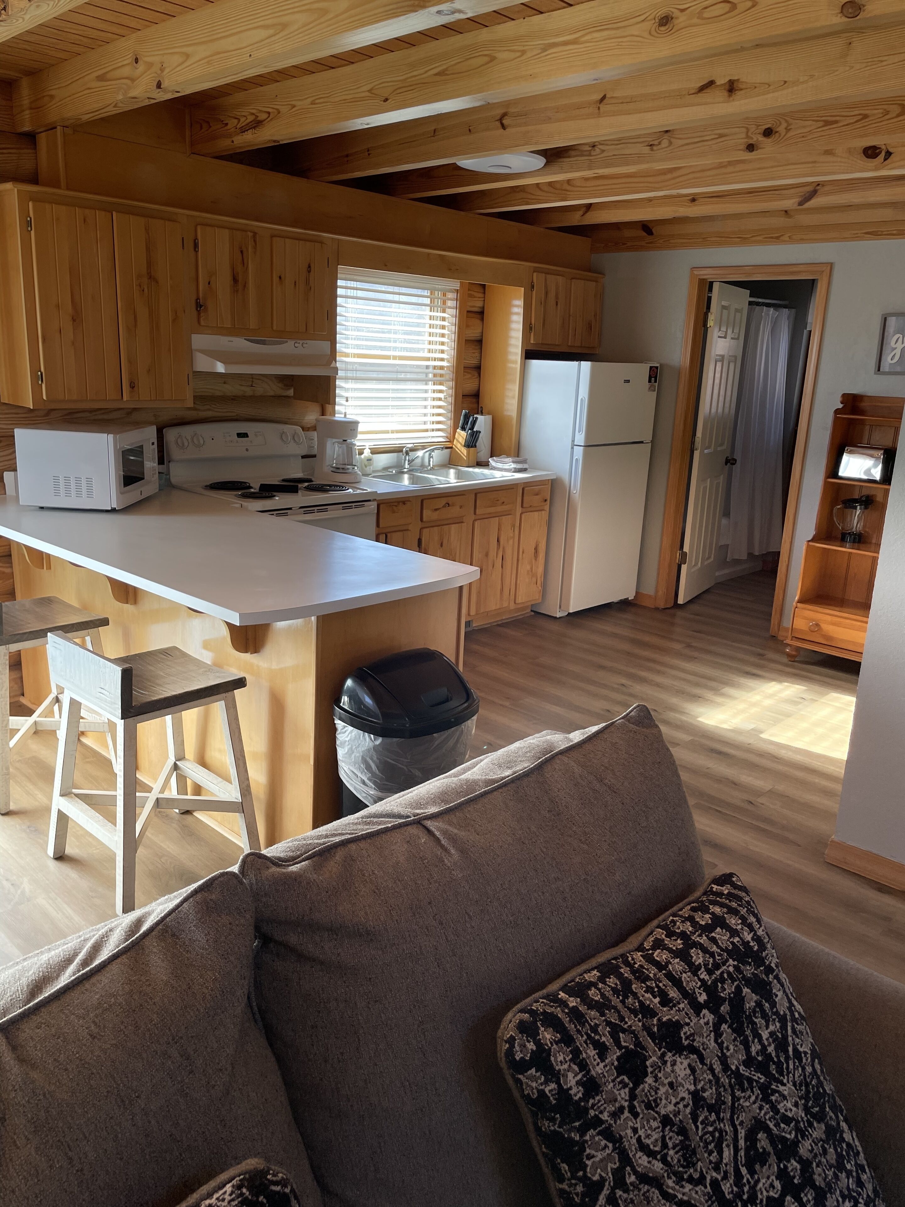 Una cocina acogedora con armarios de madera, encimera blanca, microondas, estufa y refrigerador. Hay un bote de basura y una ventana con persianas.