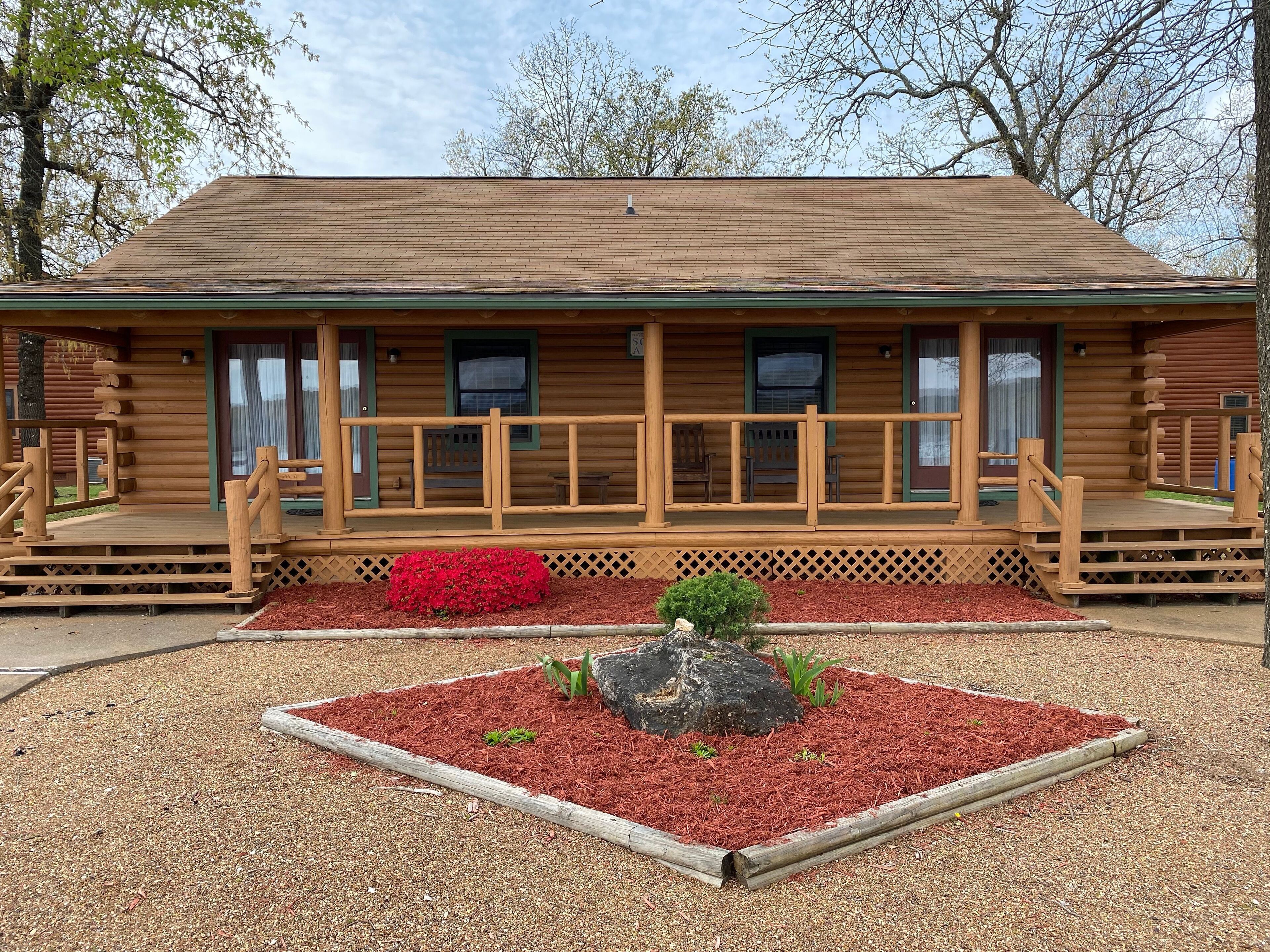 A log cabin with a porch, a small garden with red mulch, and a rock centerpiece.