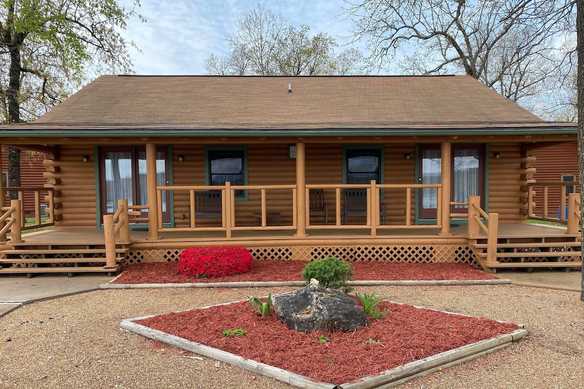 Une cabane en rondins avec un porche, un petit jardin avec un paillis rouge et un élément décoratif central en pierre.