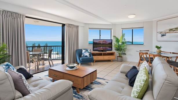Uma sala de estar moderna com uma televisão grande, assentos confortáveis, área de refeições e uma varanda com vista para o mar.