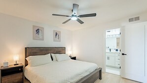 Une chambre à coucher comprenant un lit, des tables de chevet, un ventilateur de plafond et une porte donnant sur une salle de bain.