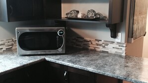 A kitchen with dark wooden cabinets, a stainless steel microwave, and a tiled backsplash. - On TheRock BnB ~ Modern Suite (Comox)