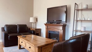 A living room with a wooden coffee table, a black leather sofa, a TV on a stand, and a fireplace. - On TheRock BnB ~ Modern Suite (Comox)