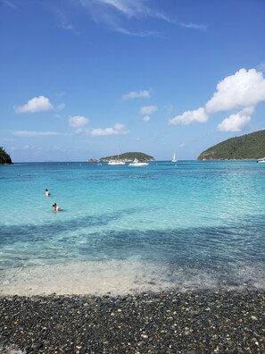 A beach with clear turquoise water, pebbly shore, and boats in the distance. - Minutes from beaches & trails of the National Park, with a large bedroom . (St.John)