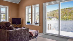 Ein Wohnzimmer mit Fernseher, einer Couch und einem großen Fenster mit Blick auf ein Gewässer und einen bewaldeten Hügel.