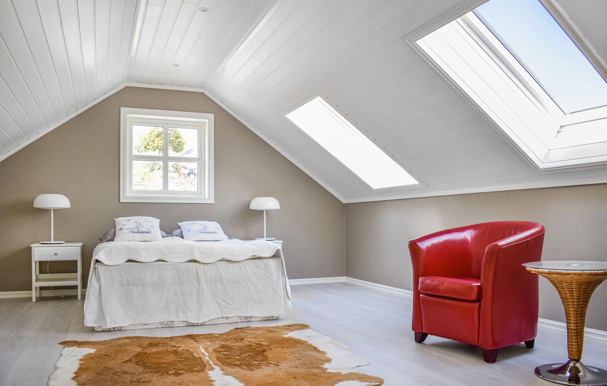 A bedroom with a sloped ceiling, a bed with white bedding, a red chair, a small table, and a cowhide rug.