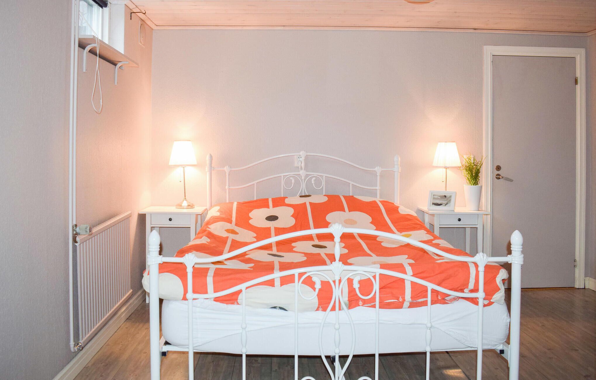 A bed with a white metal frame and an orange floral bedspread, flanked by two bedside tables with lamps, in a room with light-colored walls and a wooden floor.