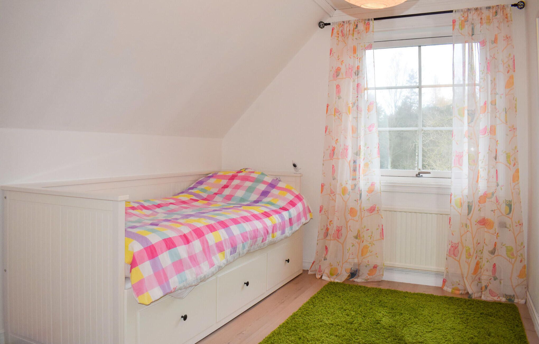 A small, single-bed room with a checkered bedspread, a window with floral curtains, and a green rug.