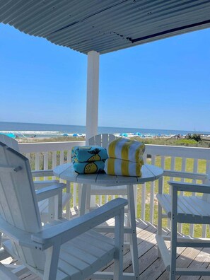 A covered outdoor seating area with white chairs and a table, overlooking a beach with umbrellas and people. - Elevator. Oceanfront. Big Deck. Pool. (Carolina Beach)