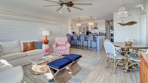 Un salon moderne avec un ventilateur de plafond, un coin repas avec des chaises en osier et une cuisine en arrière-plan.