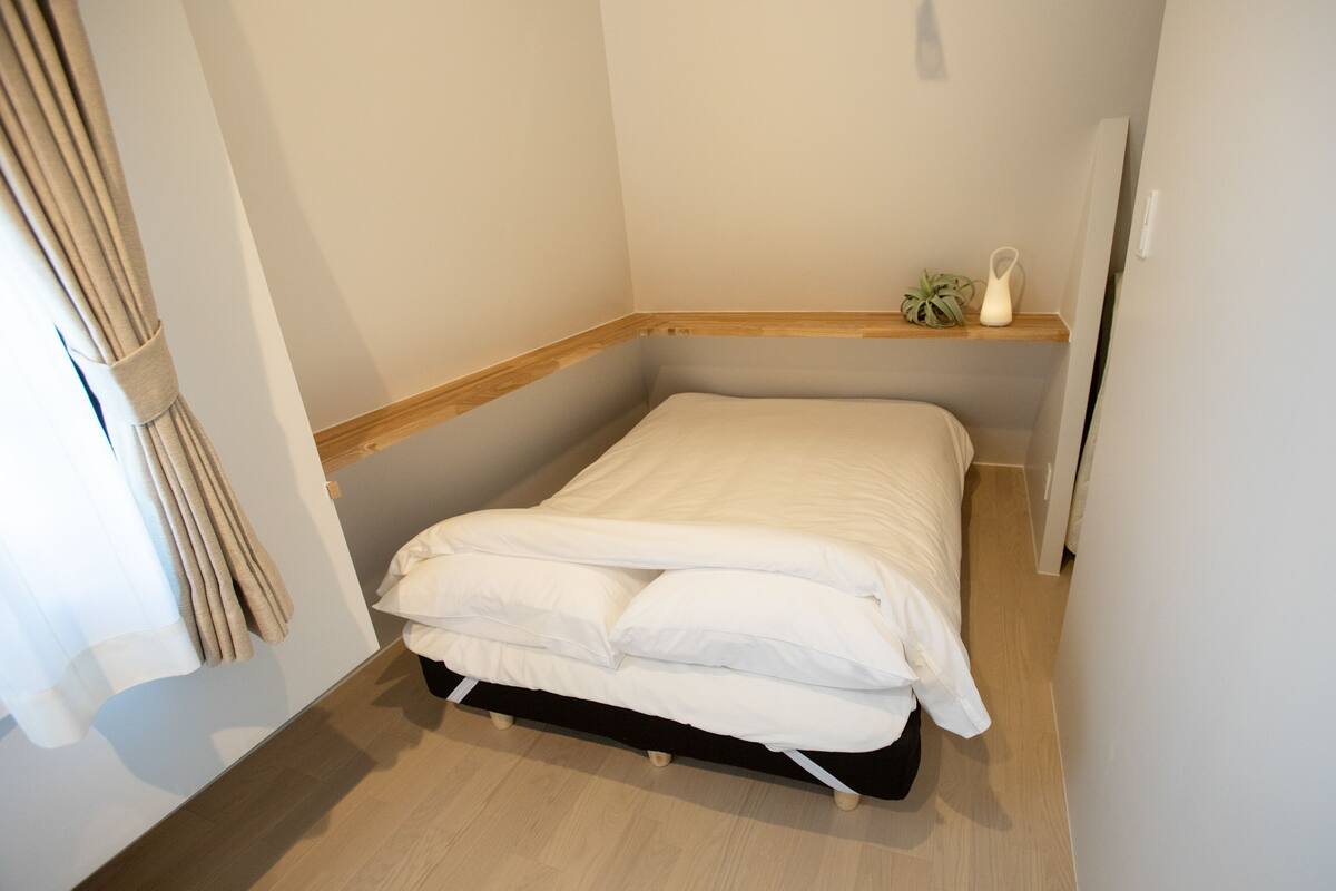 A small, neatly made bed in a corner room with a wooden floor, a small shelf with a plant, and a curtain on the window.