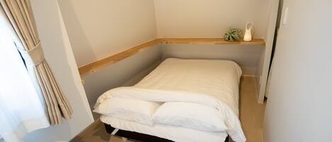 Una cama pequeña y bien hecha en una habitación de esquina con suelo de madera, una estantería pequeña con una planta y una cortina en la ventana.