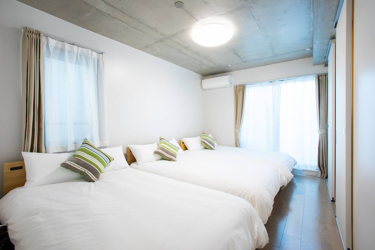 A hotel room with two beds, beige curtains, a ceiling light, and an air conditioning unit.