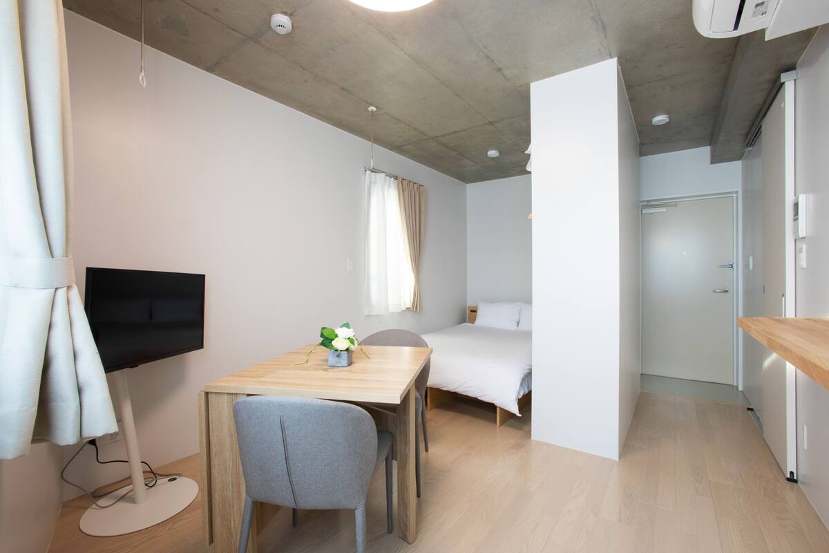 A modern, compact room with a wooden desk, a flat-screen TV, a bed, and a kitchenette.