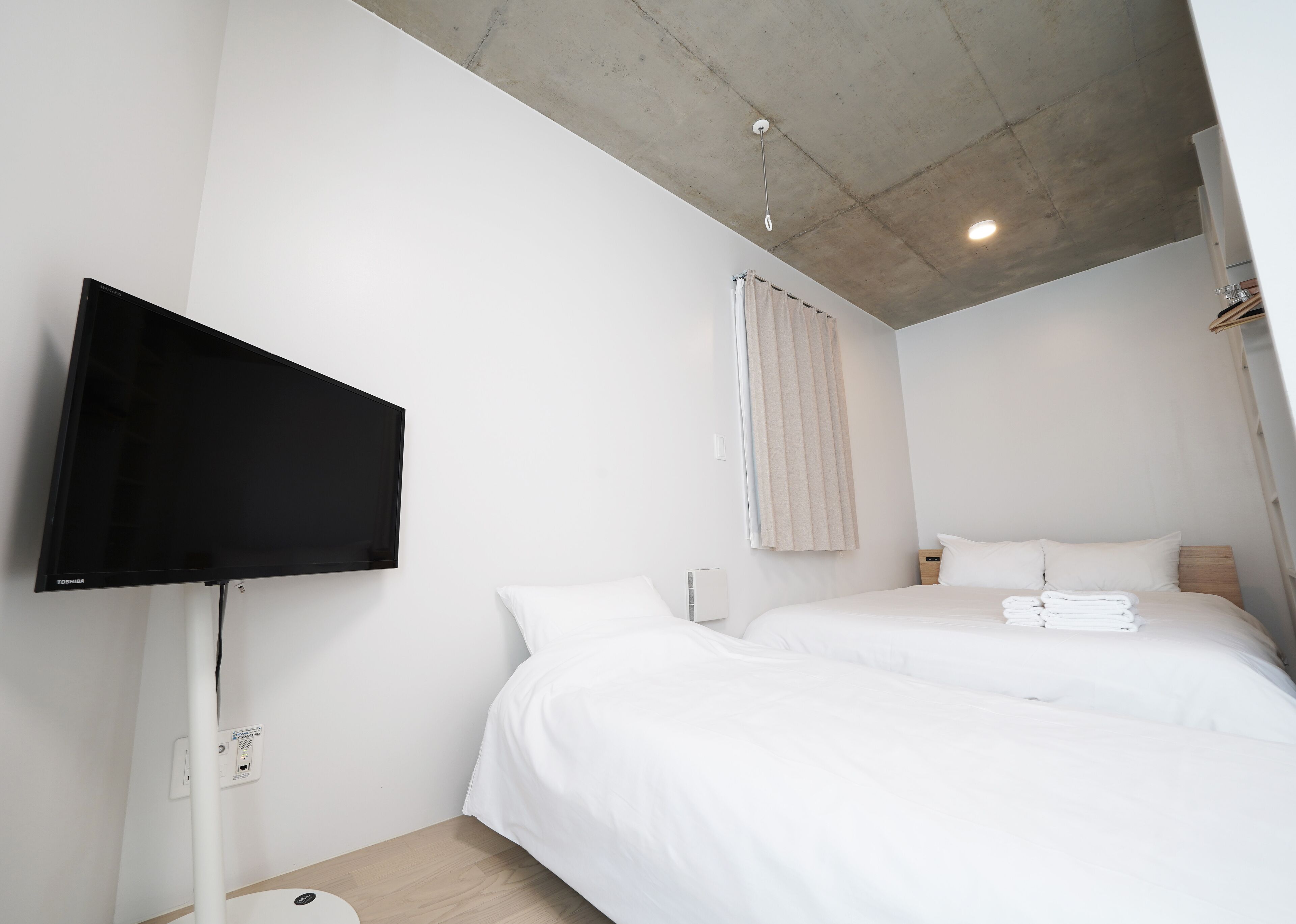A hotel room with two beds, a flat-screen TV mounted on the wall, a ceiling light, and a window with curtains.