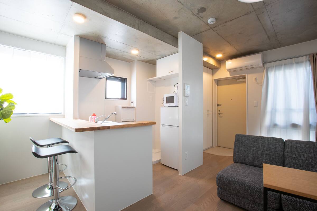 A compact kitchen with a bar stool, a microwave, and a refrigerator. There is a grey sofa and a wooden table in the living area.