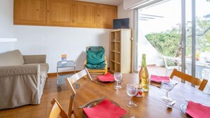 A dining area with a wooden table set for four, a sofa, a TV on a stand, and a view of greenery outside.