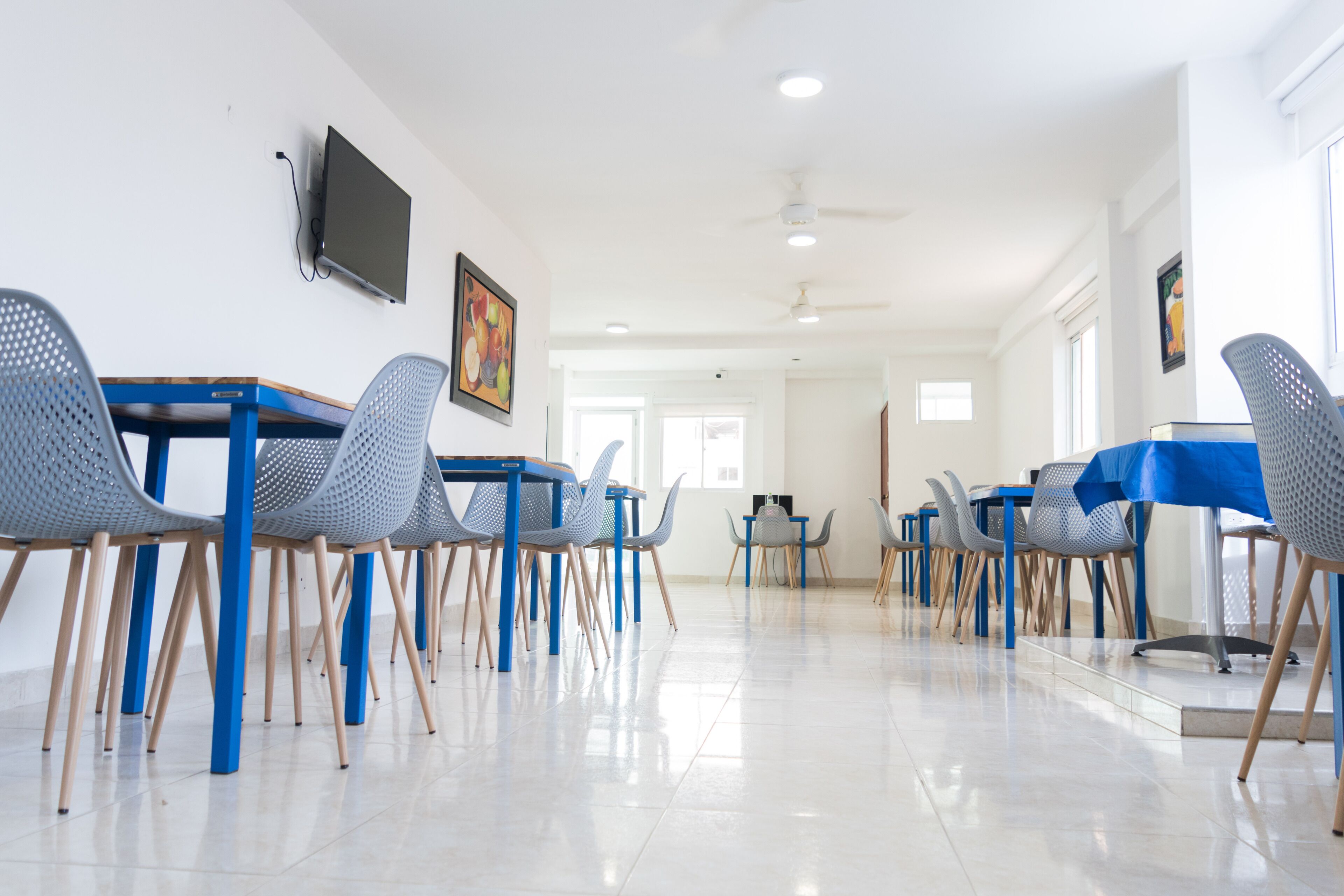 Un grand espace dédié à la salle à manger, avec des tables et des chaises bleues, un ventilateur de plafond et un téléviseur fixé au mur.