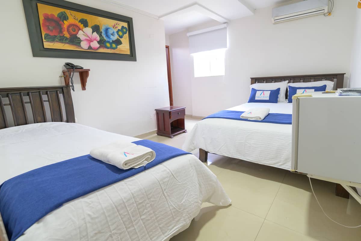 A hotel room with two beds, each with a blue bedspread and white linens, a wooden headboard, and a painting of flowers on the wall.