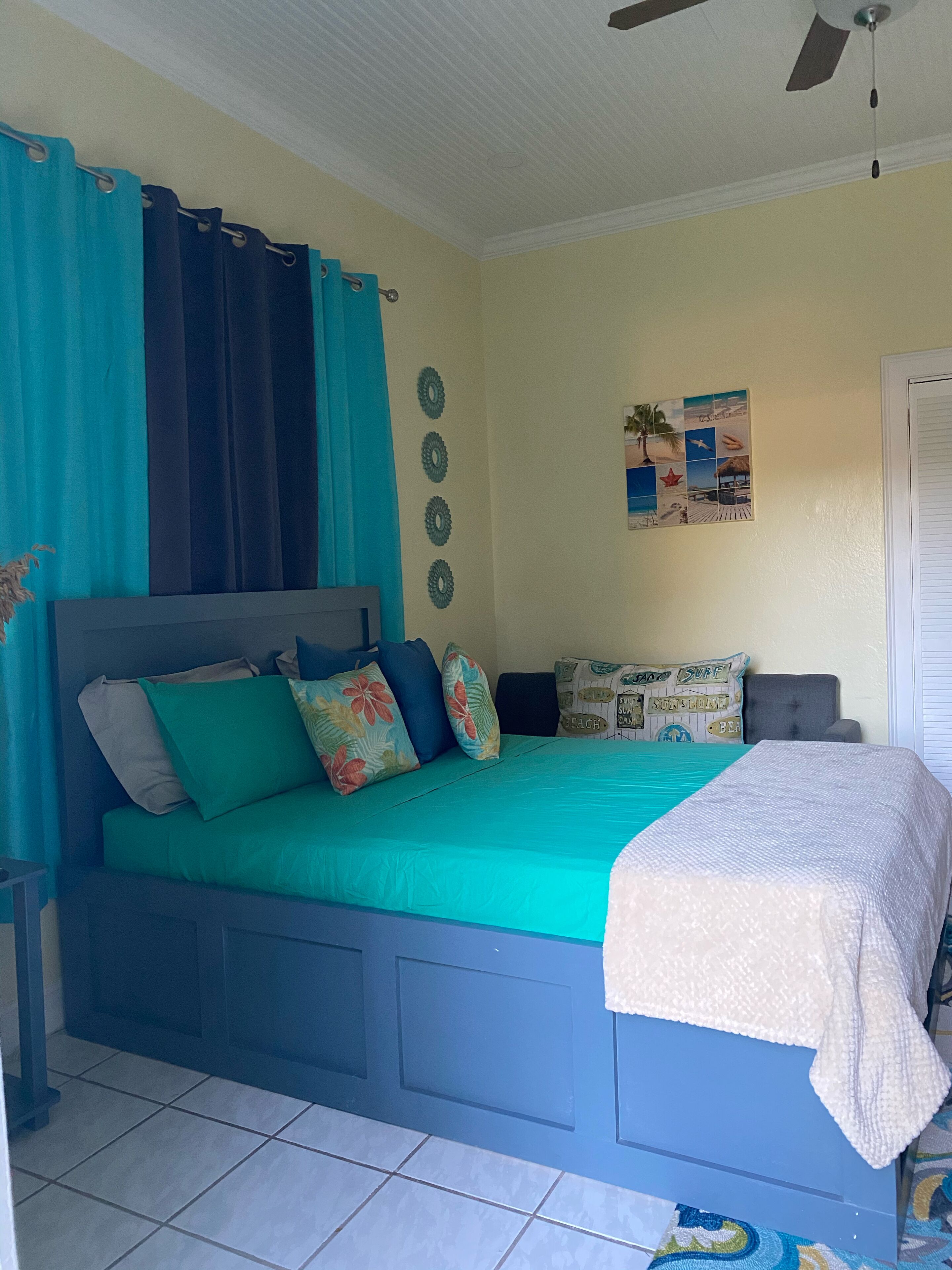 A bedroom with a bed, curtains, a ceiling fan, a wall clock, and a beach-themed pillow.