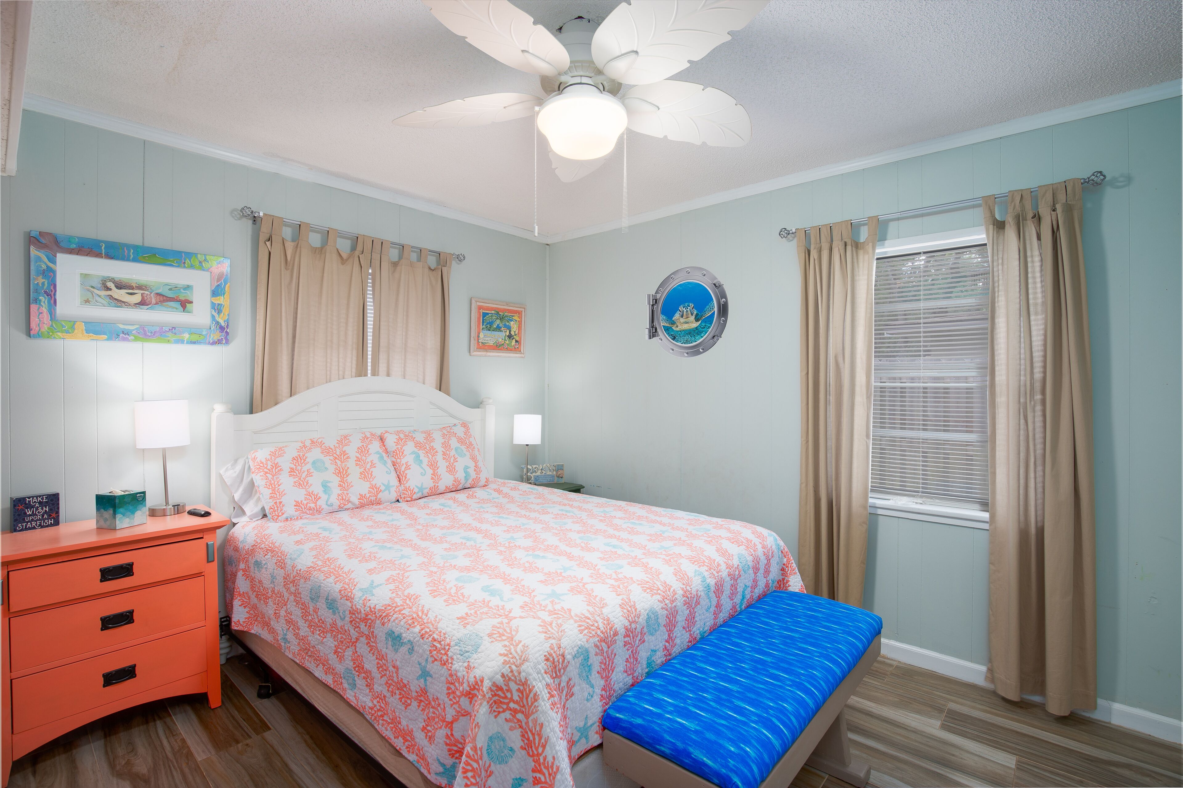 Un dormitorio con cama, mesita de noche, banco, ventilador de techo y ventana con cortinas.
