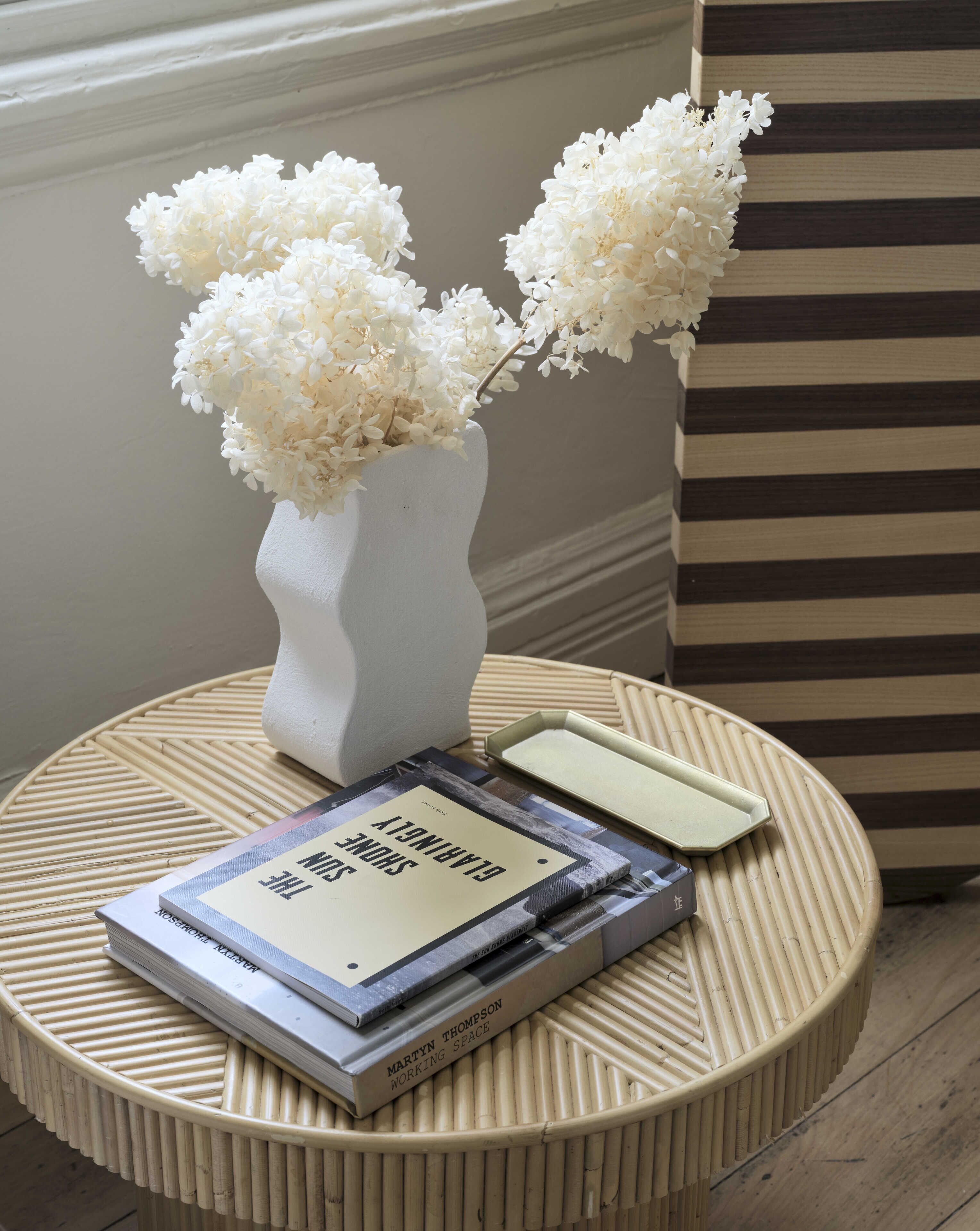A round table with a white vase containing white flowers, a book titled 'Working Space' by Martyn Thompson, and a closed book with a beige cover.