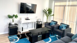 Un salon moderne avec un téléviseur à écran plat, deux plantes en pot, un canapé gris avec des coussins bleus, une table basse noire et un ventilateur de plafond.