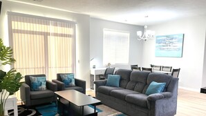 Un salon moderne avec un canapé gris, deux fauteuils, une table basse et, au fond, un coin repas.