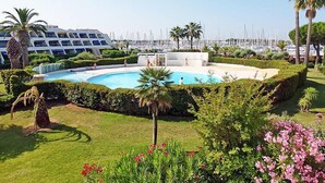 Una piscina rodeada de abundante vegetación, palmeras y un puerto deportivo con barcos al fondo.