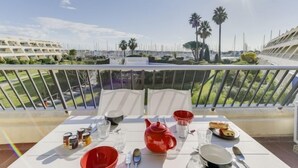 Un balcón con una mesa preparada para el desayuno, con vista a un puerto deportivo con barcos y palmeras.
