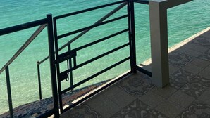 Duidelijk uitzicht op turquoise water vanaf een balkon met een metalen hekwerk, een betegelde vloer en een verre kustlijn onder een licht bewolkte lucht.
