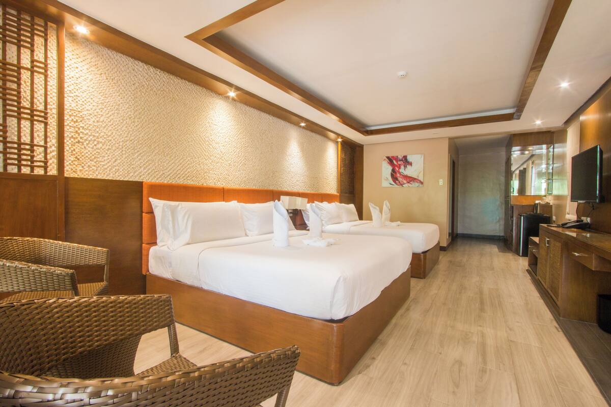 Une chambre d’hôtel avec un grand lit, deux tables de chevet, une télévision et un coin salon avec des fauteuils en osier.
