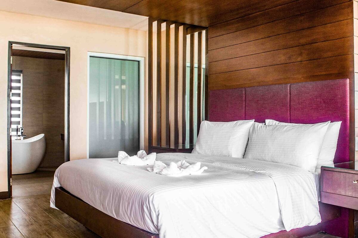 Une chambre moderne avec un plafond en bois, un grand lit recouvert de draps blancs, une table de chevet en bois et une salle de bain avec un lavabo et une baignoire blancs.