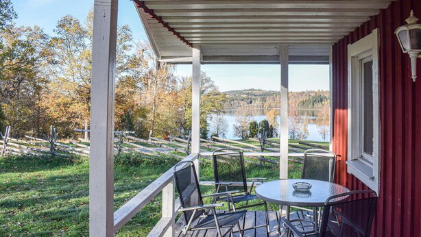 Een overdekt terras met een tafel en stoelen, met uitzicht op een meer en bebost landschap.