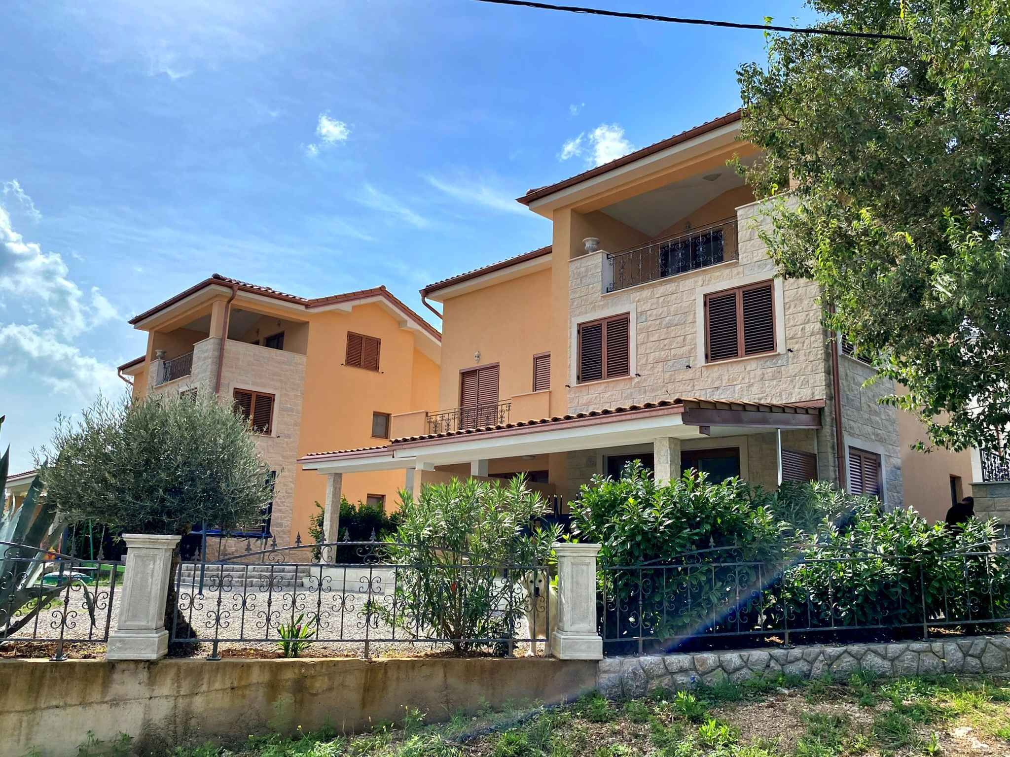 A two-story house with a balcony, a stone facade, and a surrounding garden.