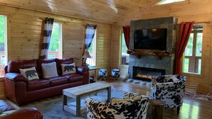 Ein gemütliches Blockhaus-Interieur mit einem Ledersofa, einem Fernseher, einem Kamin aus Stein und einer rustikalen Holzbalkendecke.
