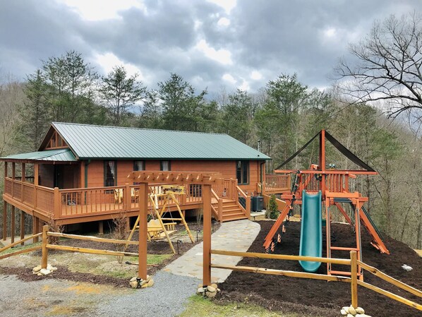 A cabin with a green roof, wooden deck, and a swing set. - Daydream Hideaway Cabin - Mtn retreat in the swaying pines, firepit, playground (Pigeon Forge)