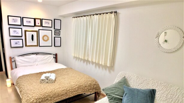 A neatly arranged bedroom with a bed, a bedside table with a candle, a white sofa with cushions, a wall with framed pictures, and a window with curtains. - Viewcity Shinjukugyoen 801-2 (Tokyo)