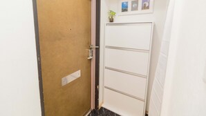 A closed wooden door with a metal handle, a white wall with a small potted plant and three framed pictures, and a white cabinet with horizontal lines. - Viewcity Shinjukugyoen 801-2 (Tokyo)