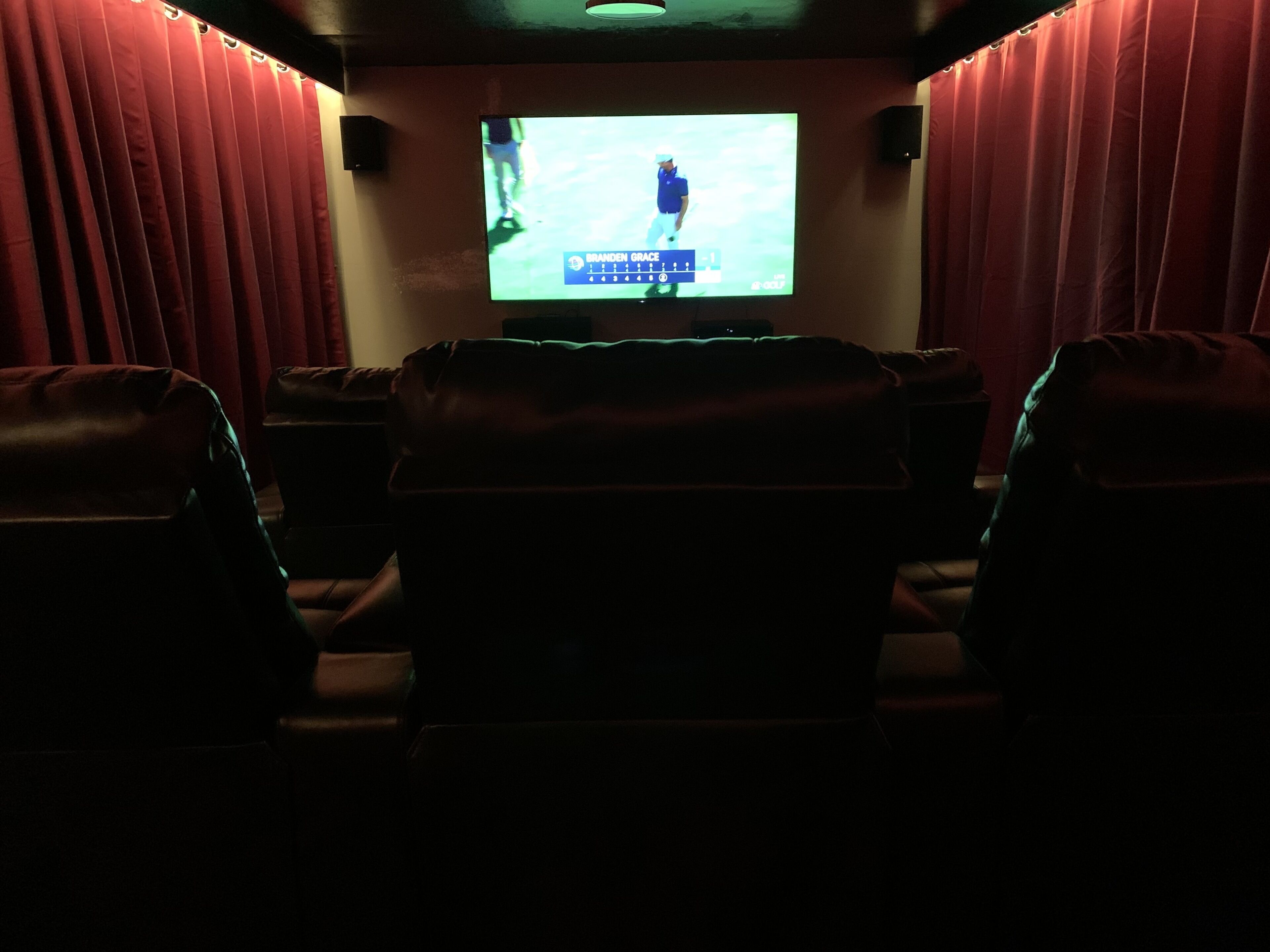 A darkened room with a large screen displaying a golf player, leather seating, and speakers on the walls.