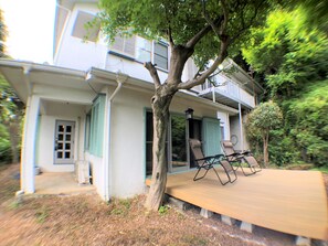 Ein zweistöckiges Haus mit Balkon, ein Baum im Vorgarten und eine Holzterrasse mit zwei Liegestühlen.