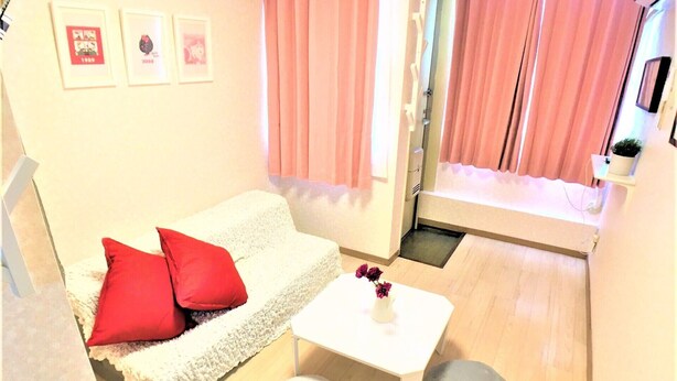 A small, cozy room with a white sofa, red cushions, a white coffee table, and two grey ottomans. There are pink curtains and a small shelf with a plant. - Bell Air OKUBO 203 (Tokyo)